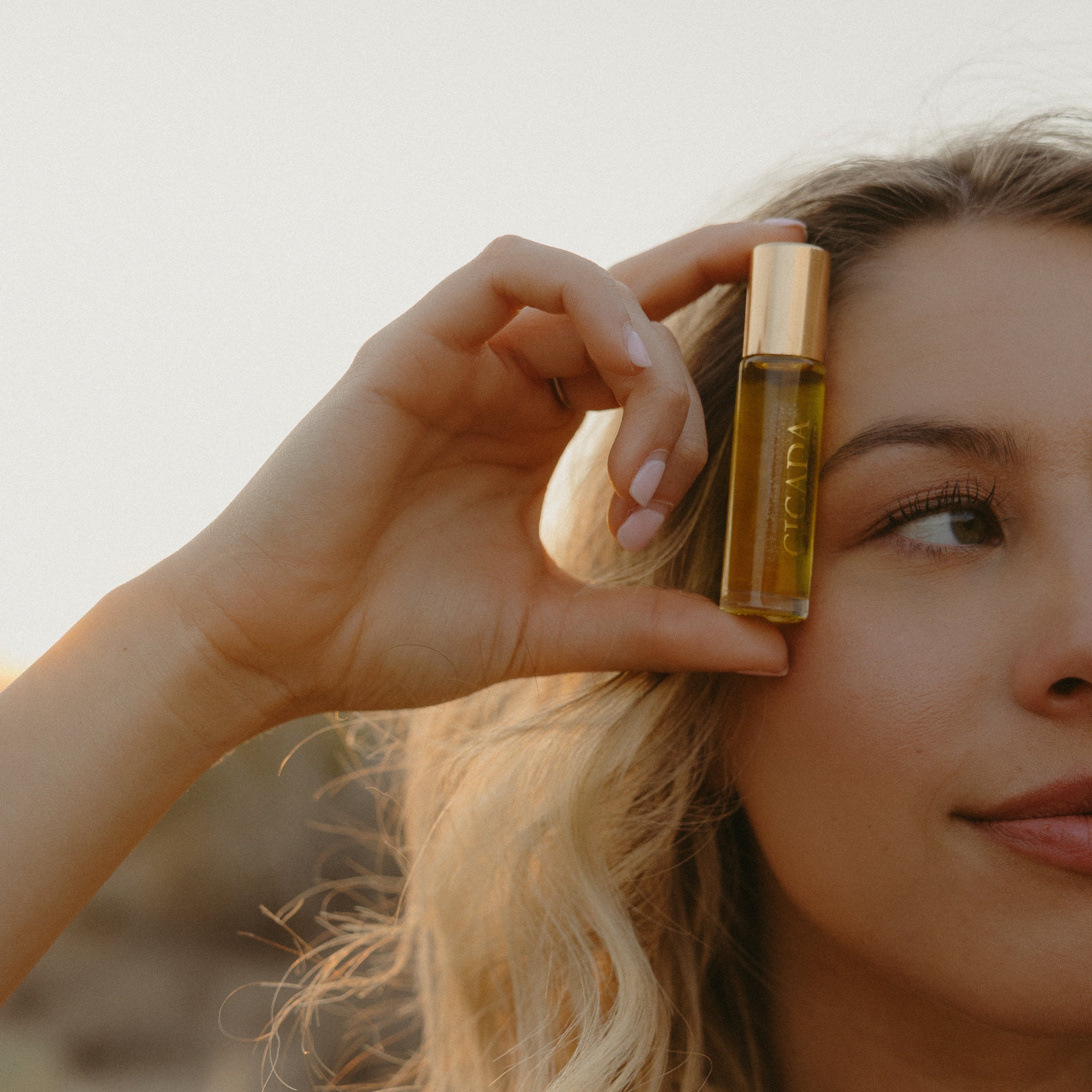 Lifestyle image of woman holding CICADA perfume oil bottle next to her face.
