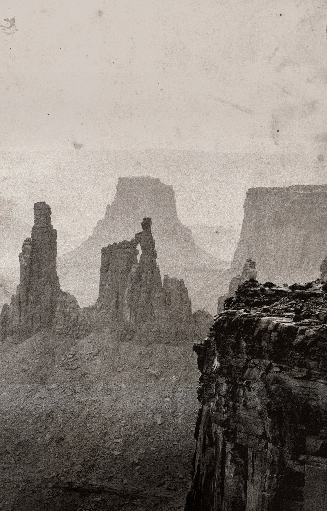 A sepia-toned landscape photograph of a rugged canyon with tall rock spires and mesas fading into mist.