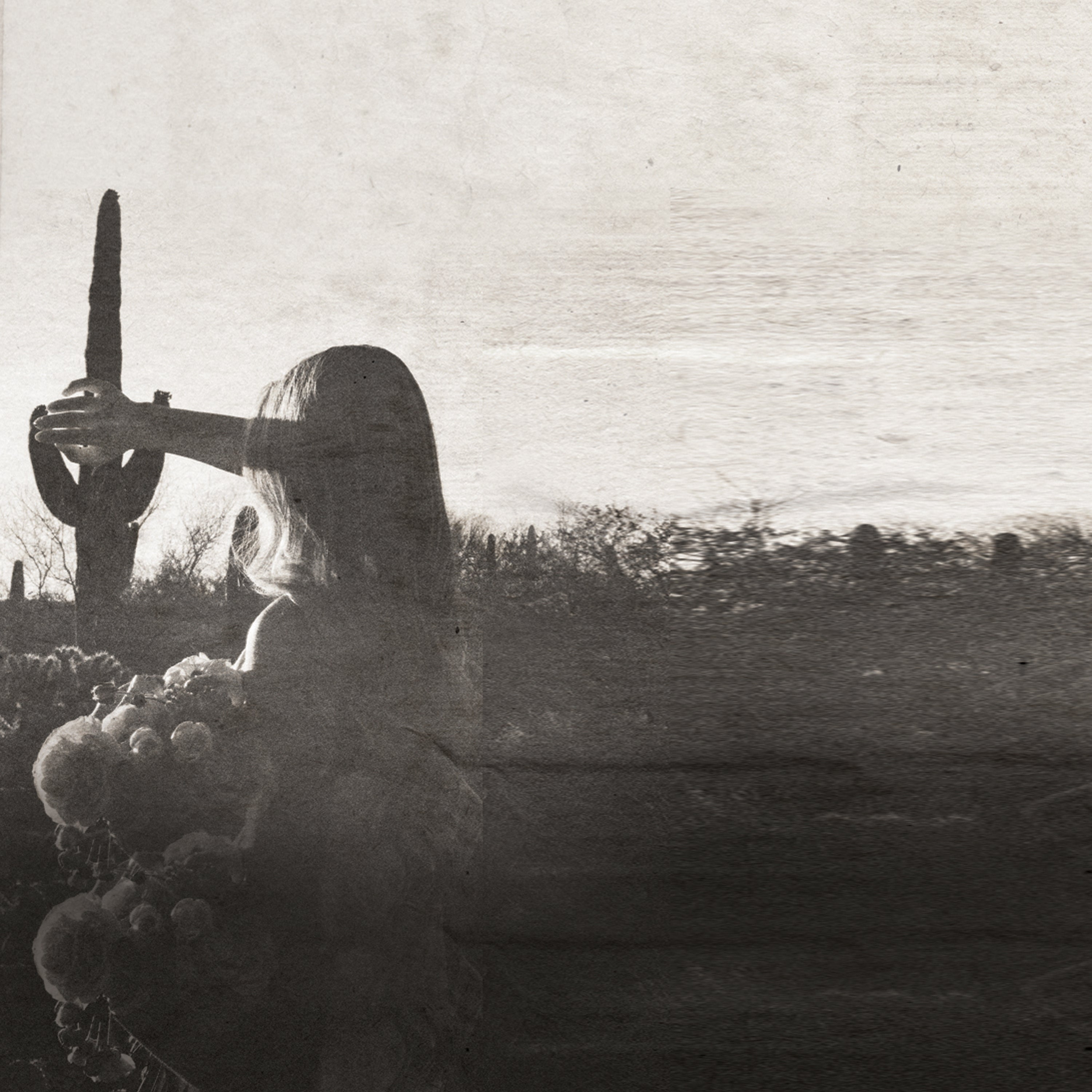 A sepia-toned artistic photograph showing a woman in profile looking at a cactus outdoors in a desert landscape, with a large blurred foreground floral shape obscuring the lower left portion of the frame.
