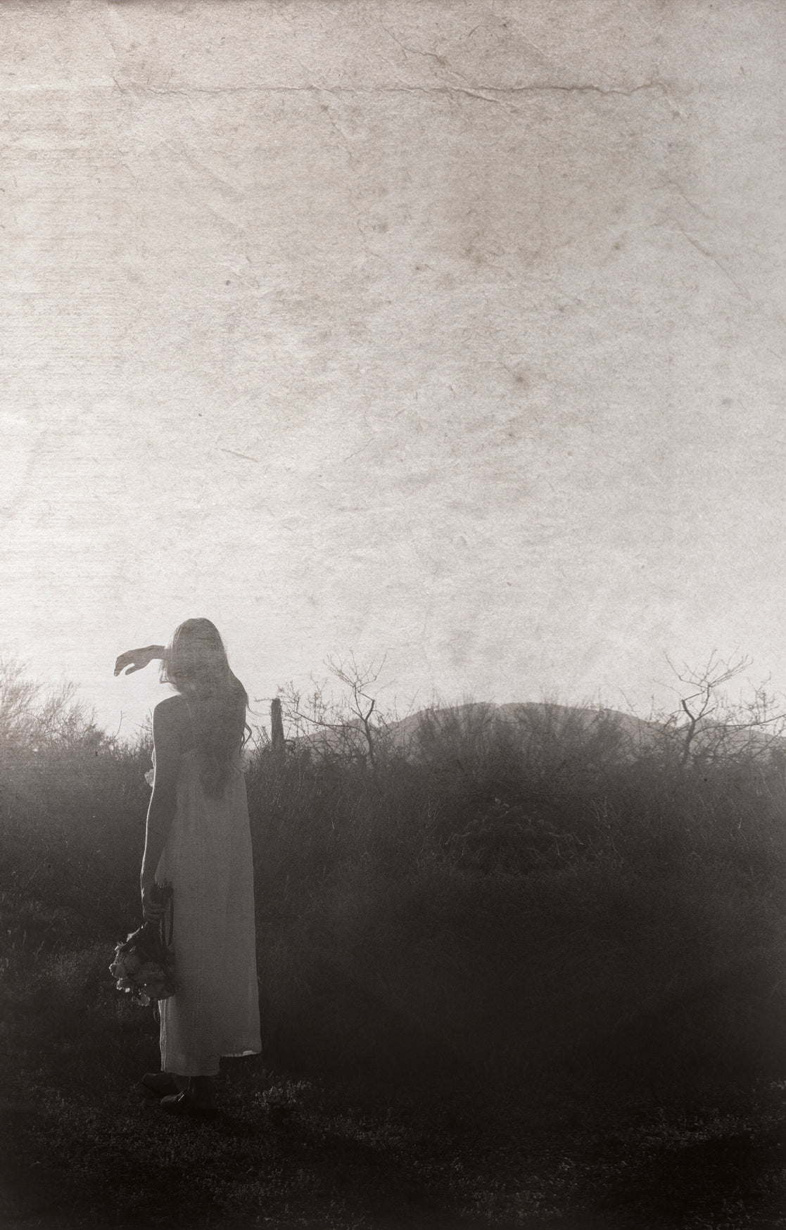 A sepia-toned artistic photograph of a woman in a long white dress standing in a dark outdoor landscape, with a large textured off-white sky area above and a hazy horizon behind her.