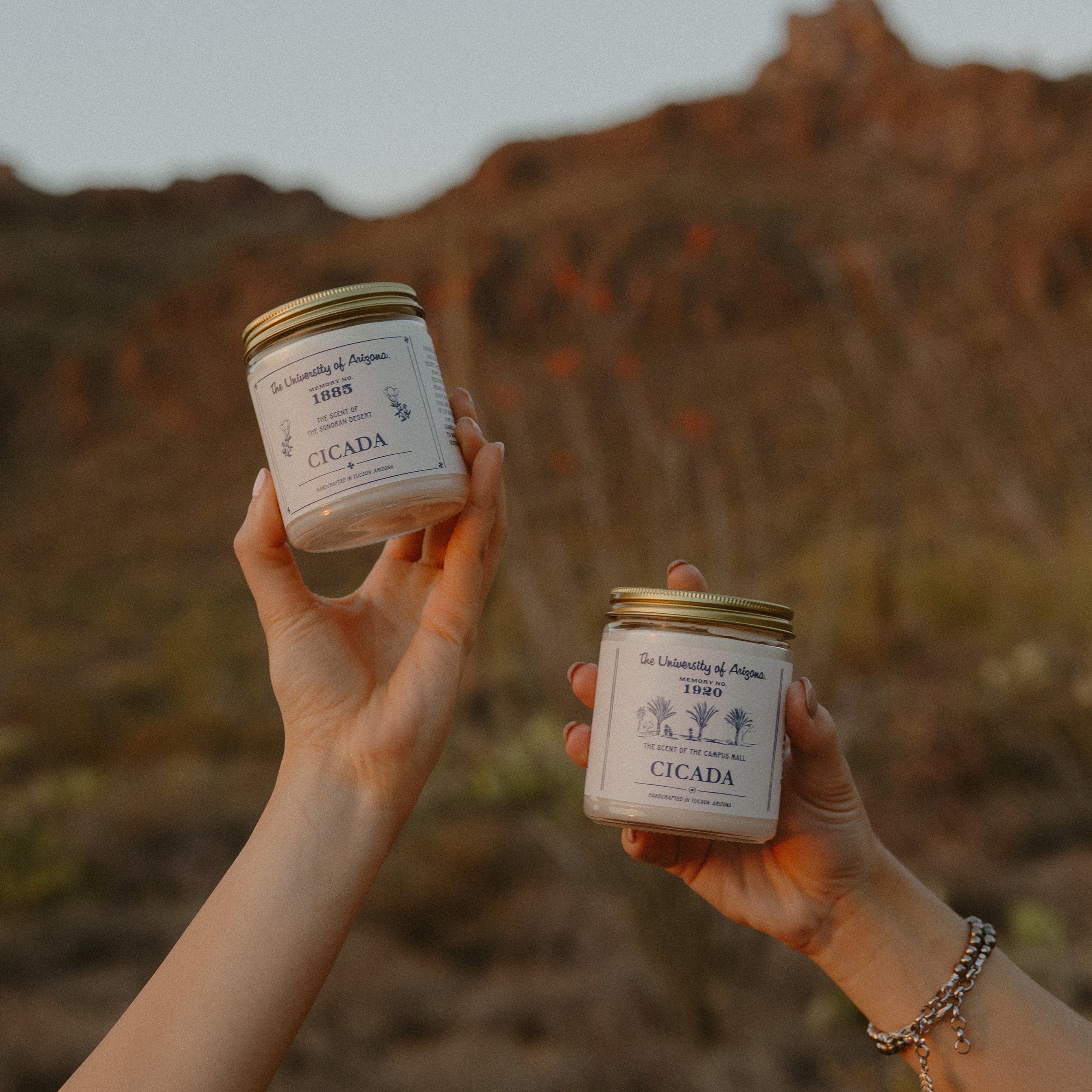Lifestyle image showing two hands holding the 7 oz glass jar candles with labels showing 'University of Arizona' and 'CICADA' logos on them, in an outdoor setting with natural landscape in the background.