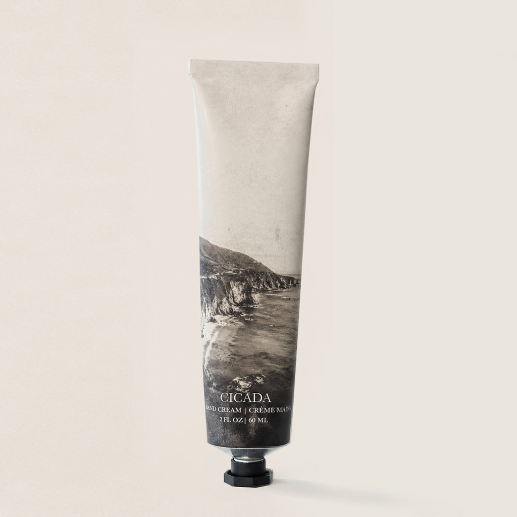 A aluminum tube of Cicada Hand Cream standing upright against a plain, warm off-white background. The tube is a textured light beige with a black and white photograph of a coastal cliffside on the lower half, and a black cap at the bottom.