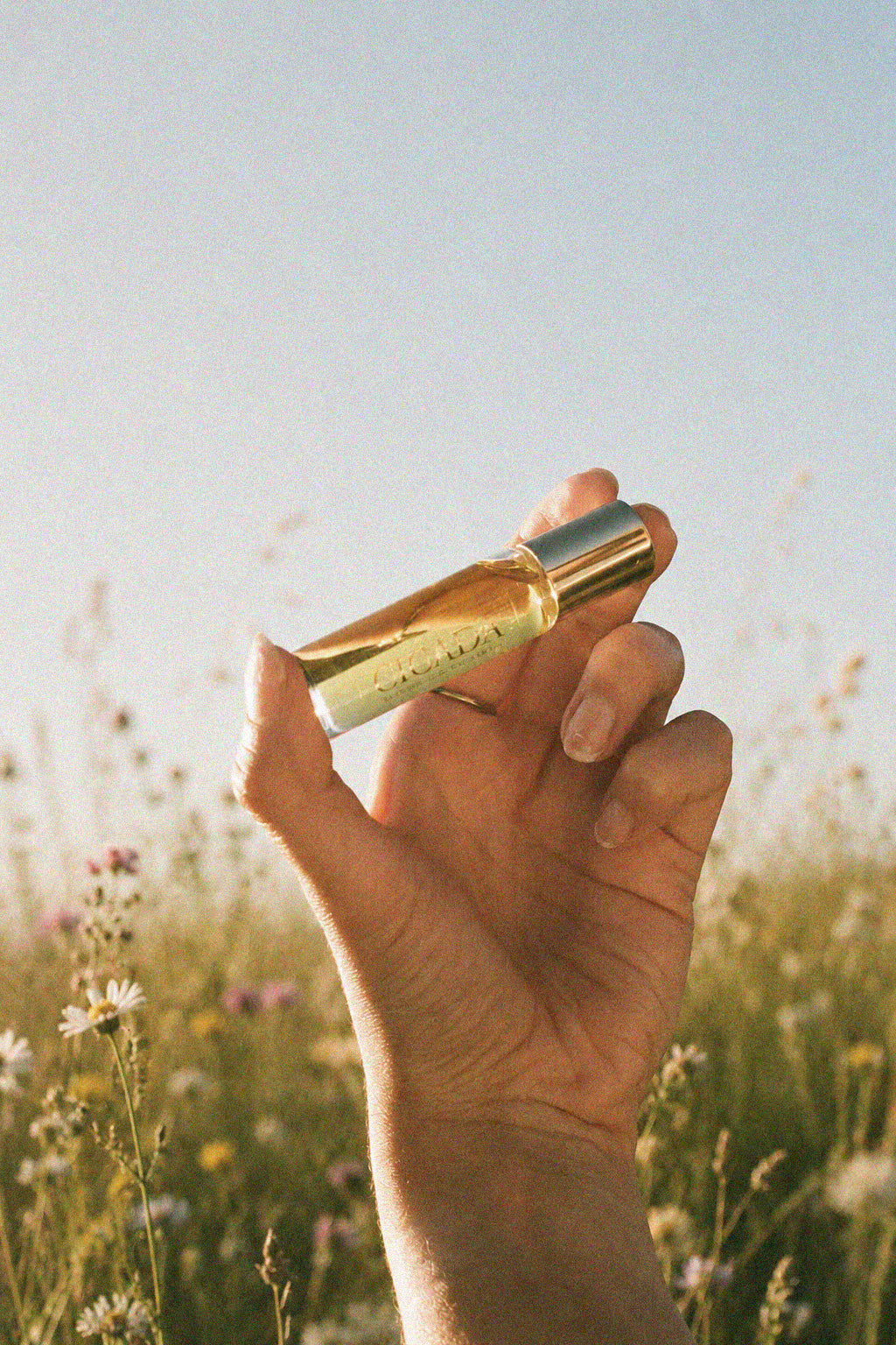 A hand holding a small gold perfume rollerball bottle with a gold cap, set against a blurred outdoor meadow background under a clear blue sky.