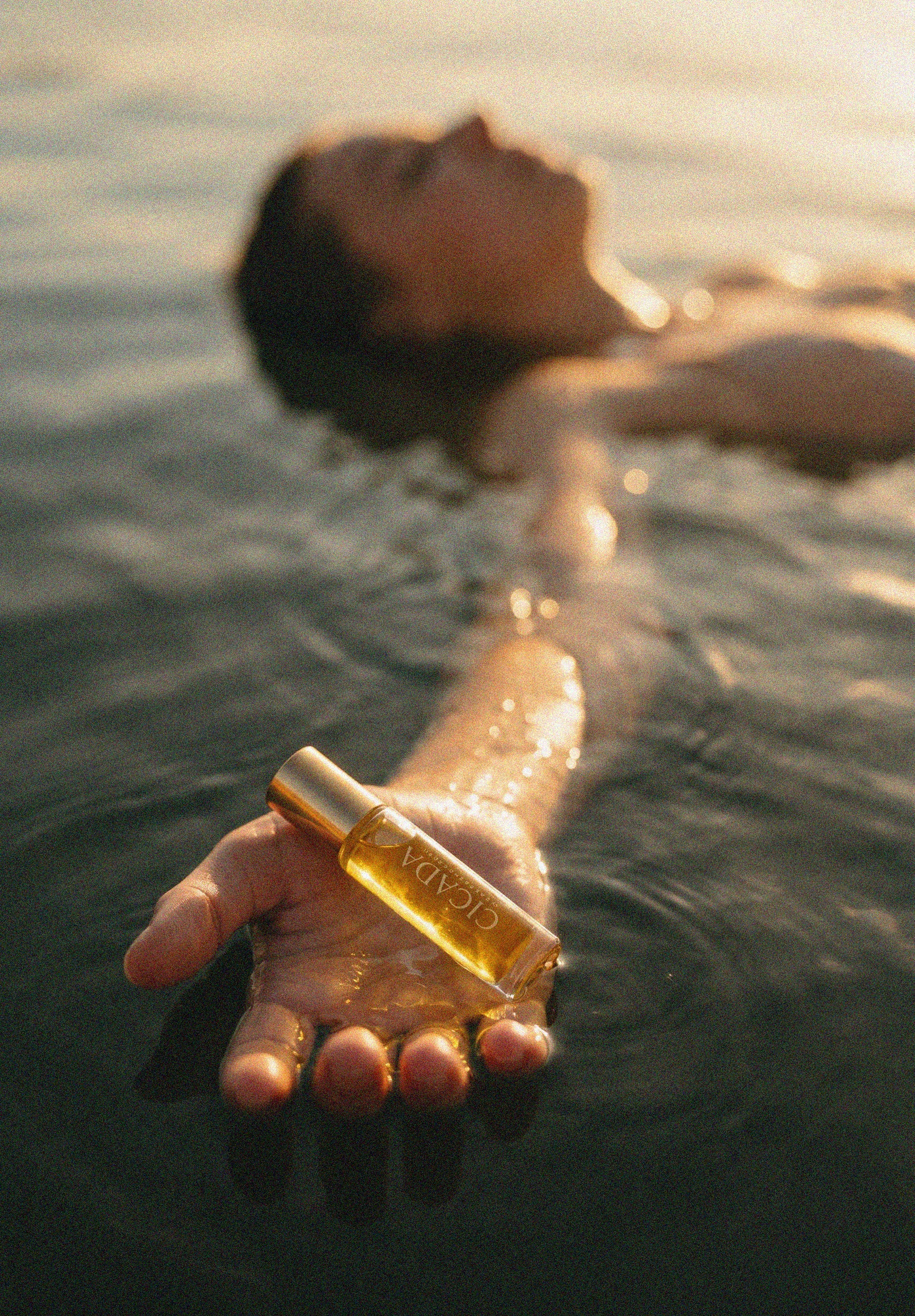 A person floating in dark water at sunset holds a small CICADA perfume oil bottle in their hand. The bottle has a gold cap and a clear body showing a golden-yellow perfume oil inside.