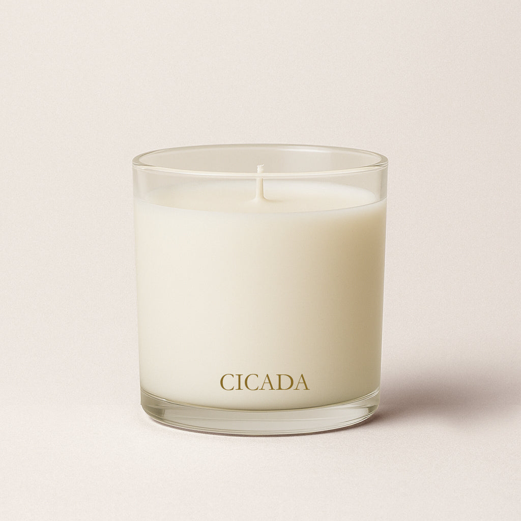 Product image of 9 oz glass vessel candle, off white colored wax, with gold brand name 'CICADA' written on it. Beige background in image. Candle is approximately 3.5