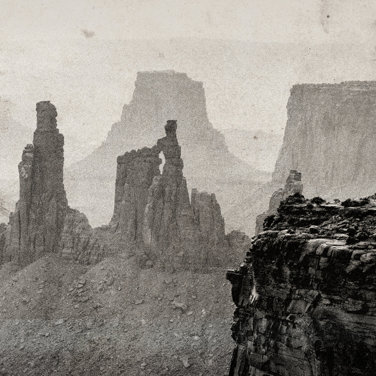 A sepia-toned landscape photograph of rugged rock formations and mesas in a canyon, with a textured, vintage print appearance.
