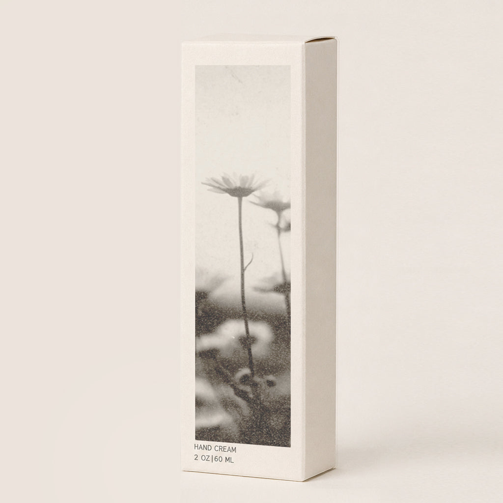 A tall, narrow cream-colored hand cream box, featuring a black-and-white photograph of daisy-like flowers on the front, set against a plain off-white background.