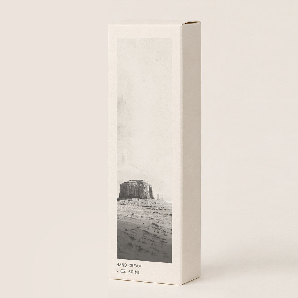 A tall, cream-colored hand cream box with a black-and-white desert landscape photo on the front, photographed on a warm off-white background.