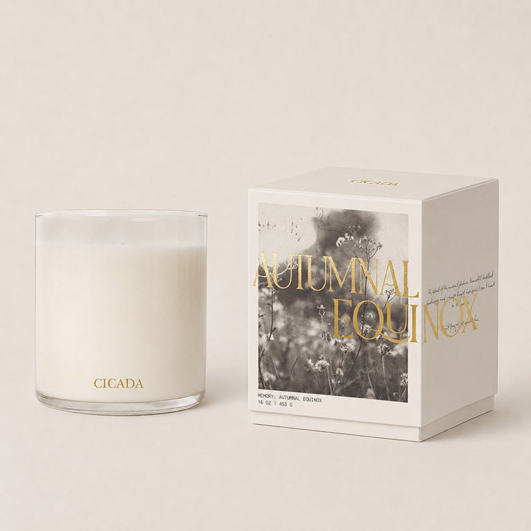 A Cicada 'Autumn Equinox' coconut-soy candle in a clear glass jar next to its cream packaging box with gold lettering and a black-and-white botanical print, set against a warm beige background.