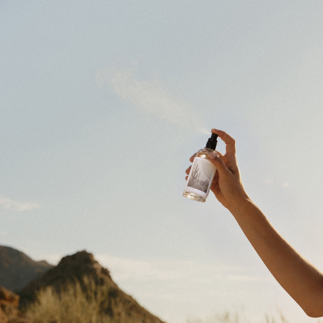 A hand holding a clear spray bottle with a black pump, spraying a fine mist into the air outdoors against a pale blue sky and blurred hills.