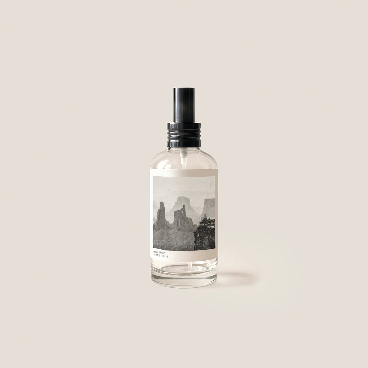 A clear glass spray bottle with a black pump dispenser and a minimalist label depicting a black and white desert rock landscape, set against a solid beige background.