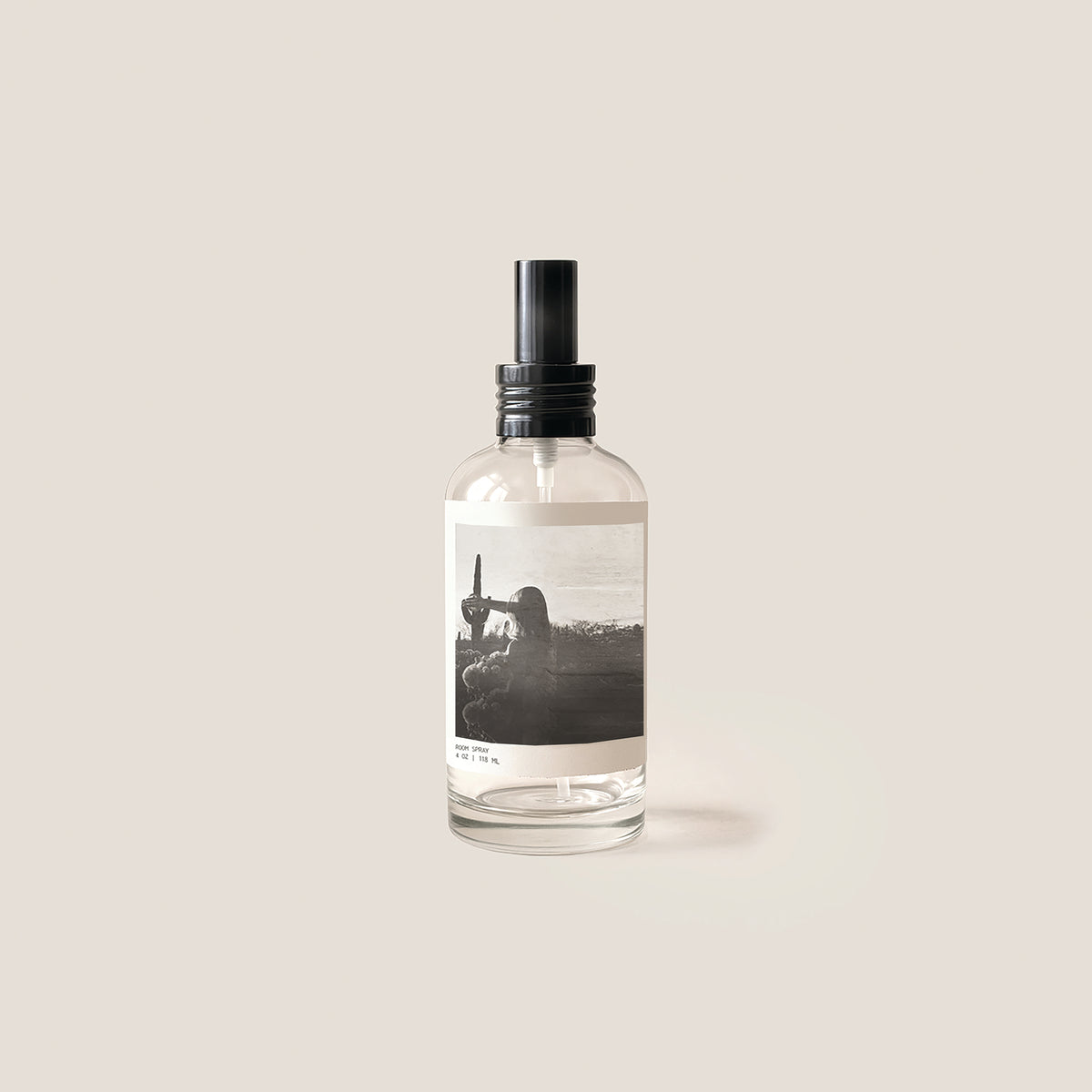 A clear glass spray bottle with a black pump dispenser and a minimalist black-and-white photographic label, centered on a solid light beige background.