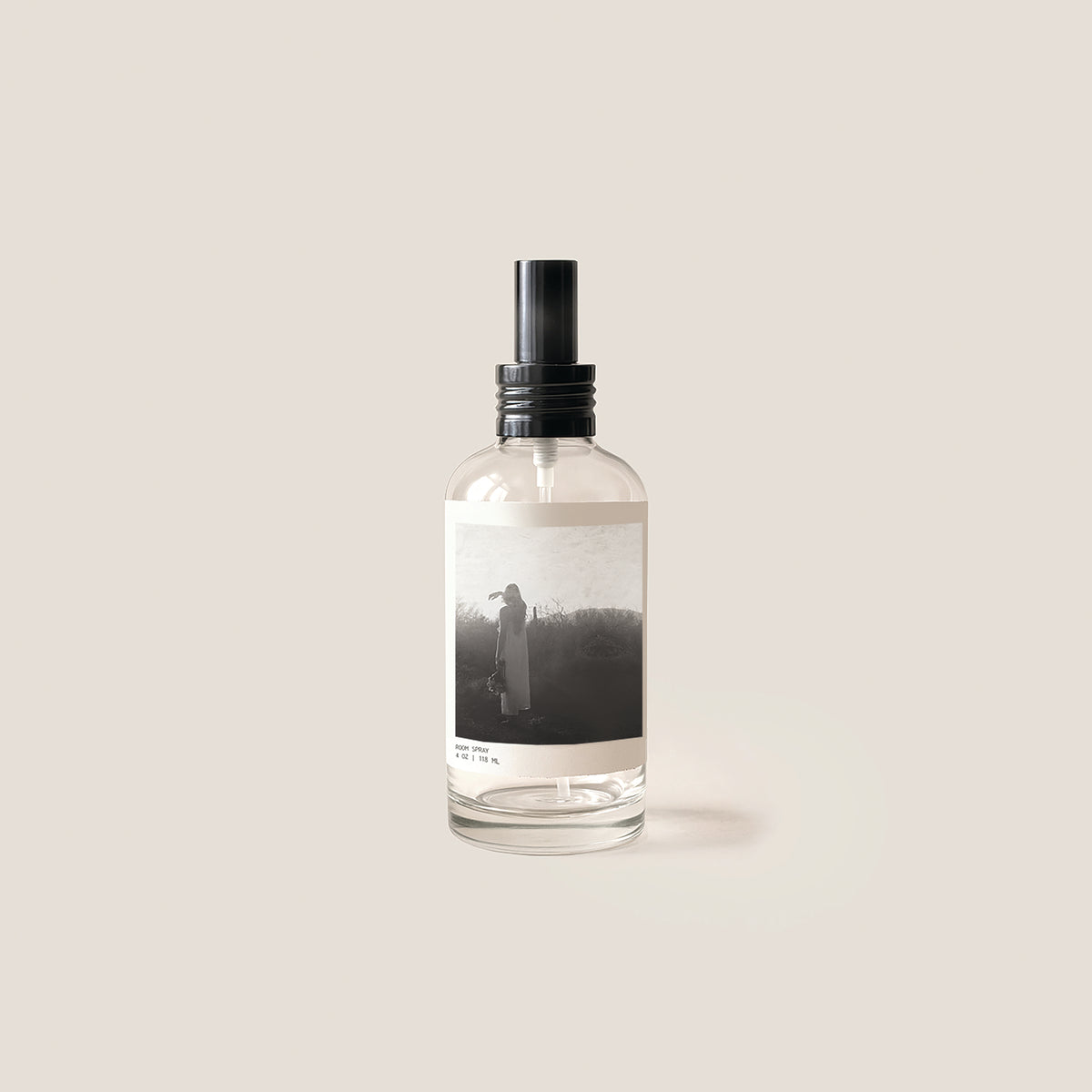 A clear glass spray bottle with a black spray top and a minimalist black-and-white label, centered on a warm beige background.