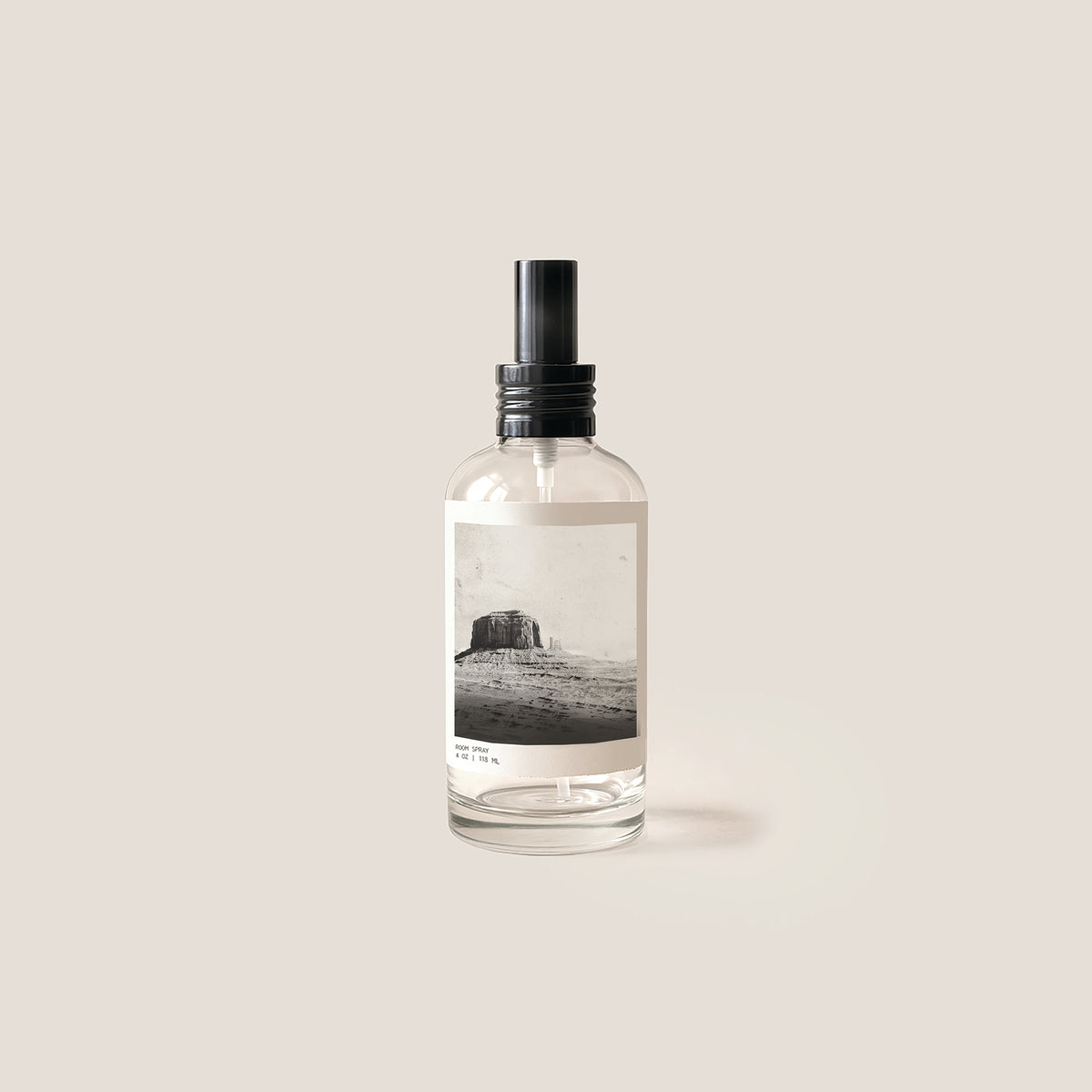 A clear glass spray bottle with a black pump dispenser and a minimalist black-and-white landscape label, centered on a warm beige background.
