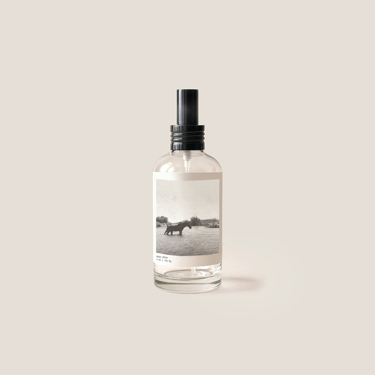 A clear glass spray bottle with a black pump dispenser and a minimalist black-and-white landscape label, centered on a warm beige background.