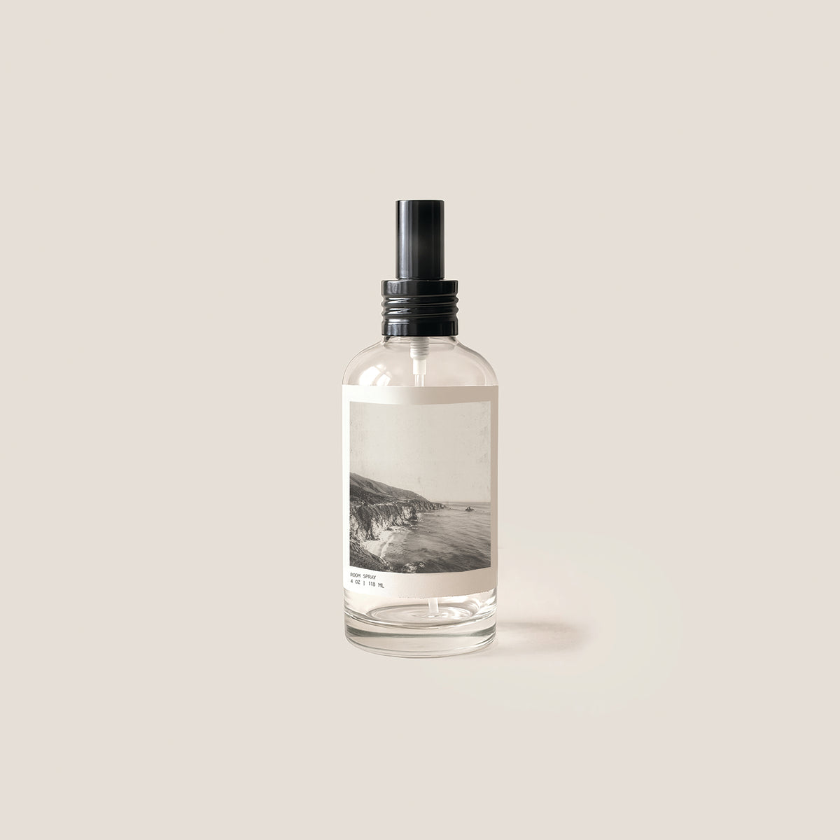 A clear glass spray bottle with a black pump dispenser and a minimalist black-and-white landscape photograph printed on the front label, set against a solid light beige background.