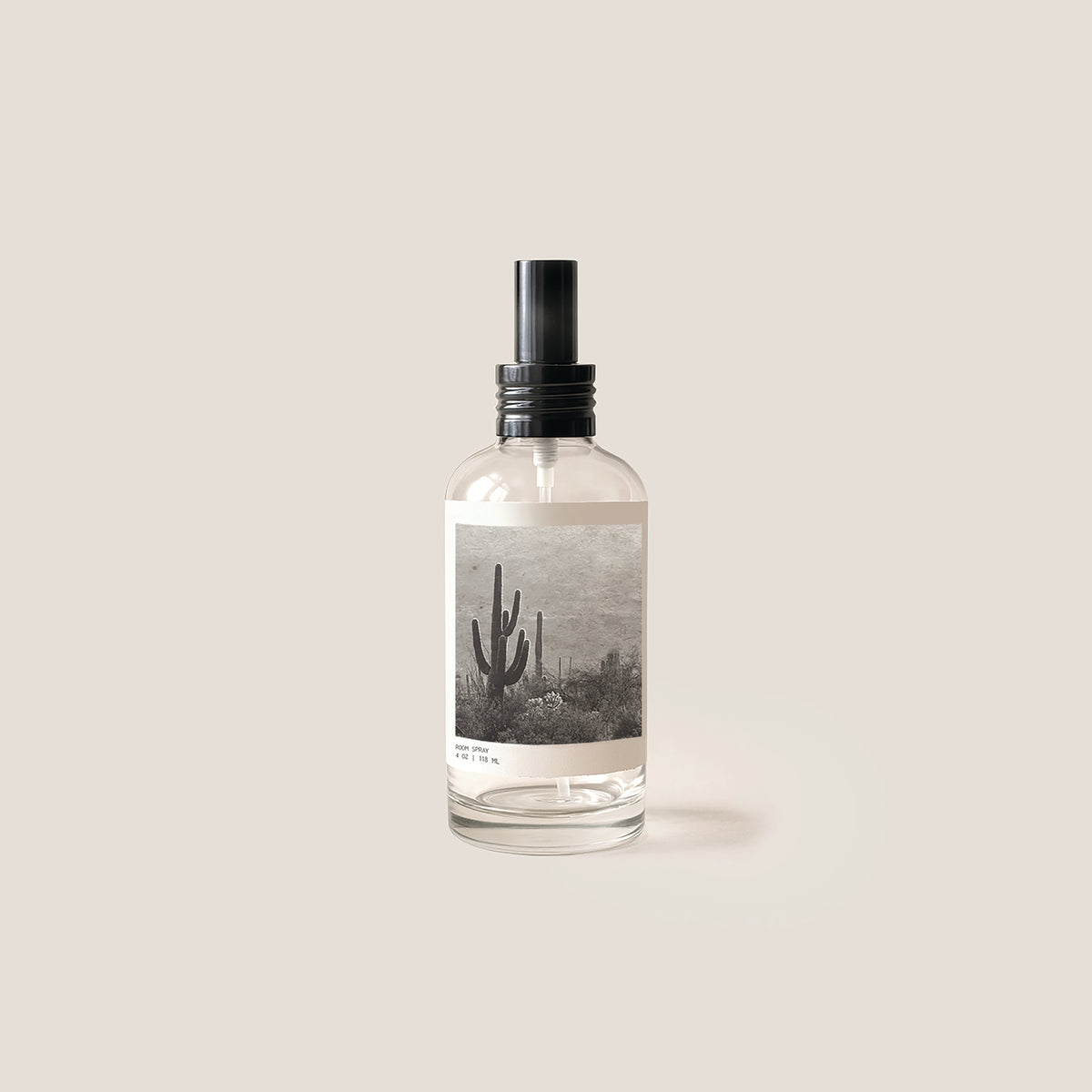 A clear glass spray bottle with a black spray top and a rectangular label featuring a black and white desert landscape with a saguaro cactus, set against a solid light beige background.