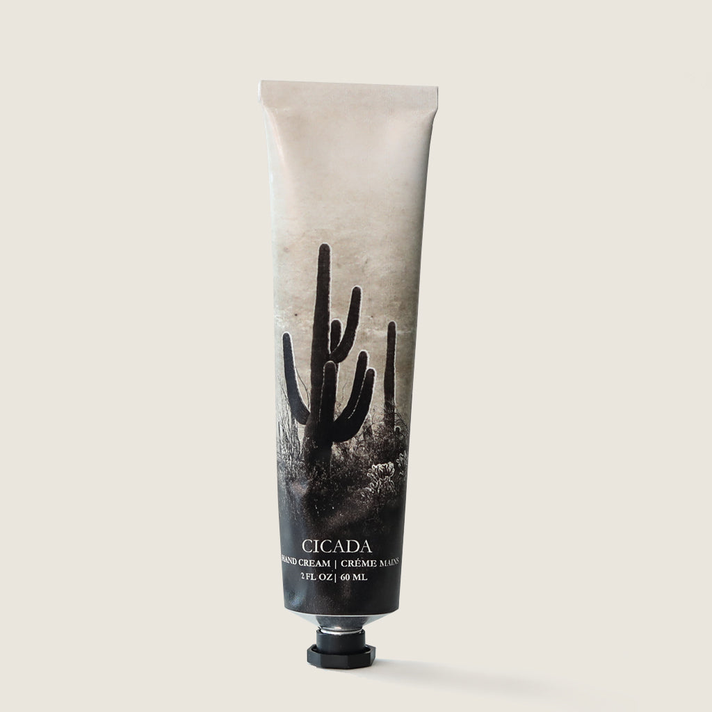 Product image of CICADA hand cream. CICADA hand cream is in aluminum tube with black and white cactus and desert landscape image on it. Hand cream tube also shows CICADA logo and the test 'Hand Cream' in both English and French. Hand cream tube also has product weight listed as '2 oz | 60 ml'. Image has cream colored background.