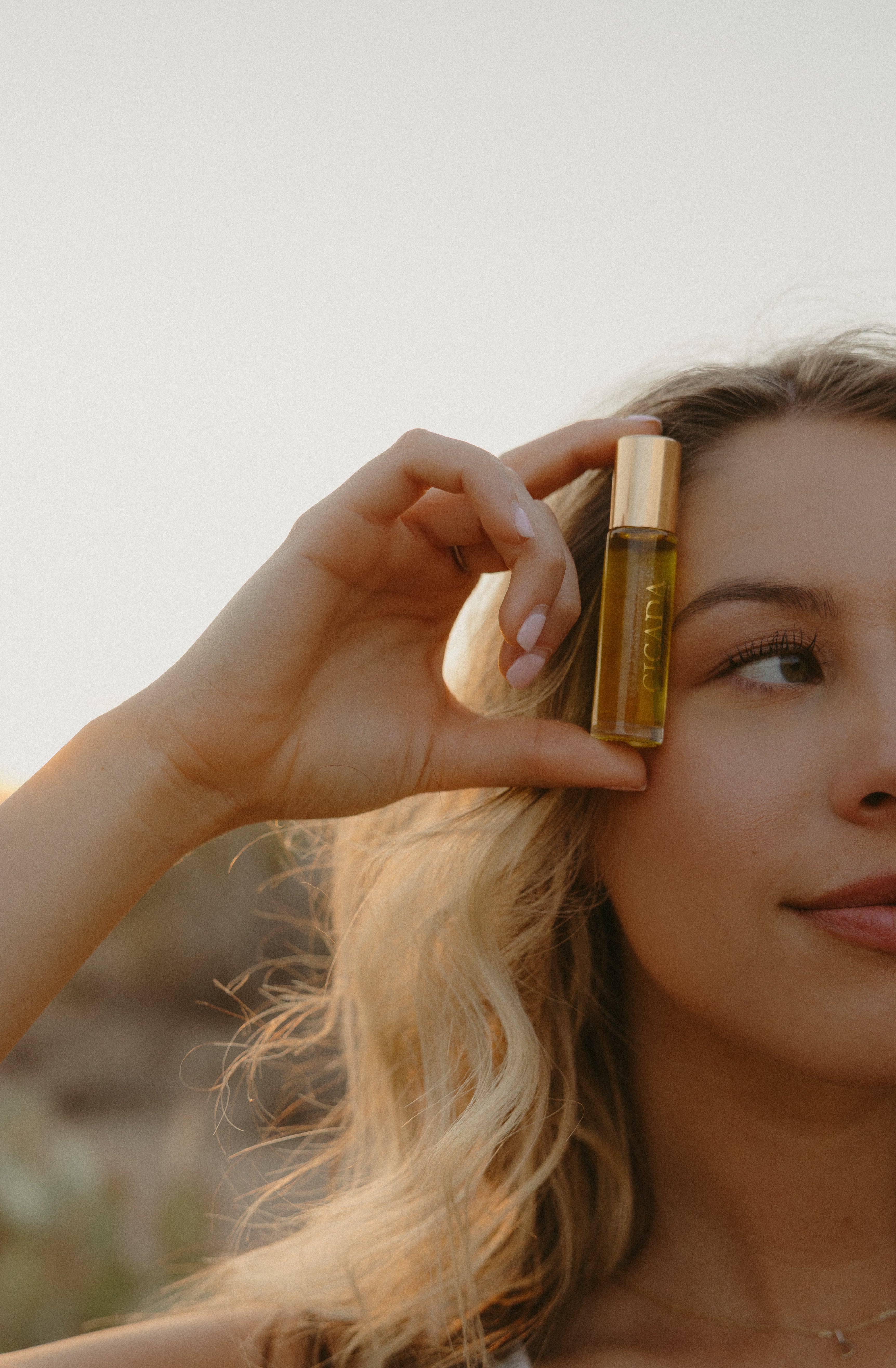 Lifestyle image of woman holding CICADA perfume oil bottle next to her face. 