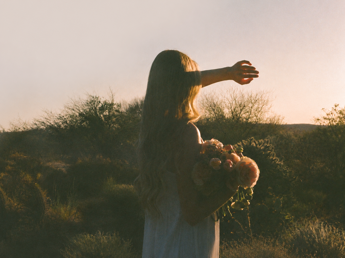 A woman with long light brown hair stands outdoors in a field at sunset, seen from behind with one arm extended upward while holding a bouquet of flowers. She is wearing a light-colored dress.