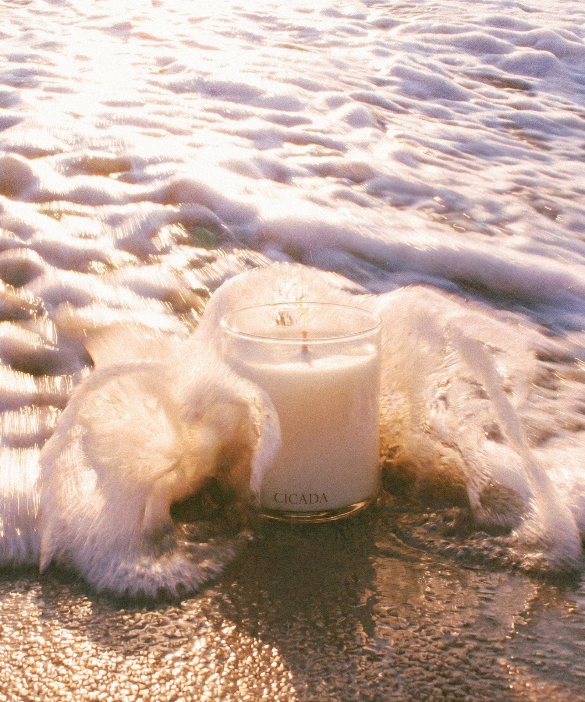 A white candle in a clear glass jar sits on wet sand surrounded by foamy ocean water, with the brand name CICADA visible near the base of the glass.