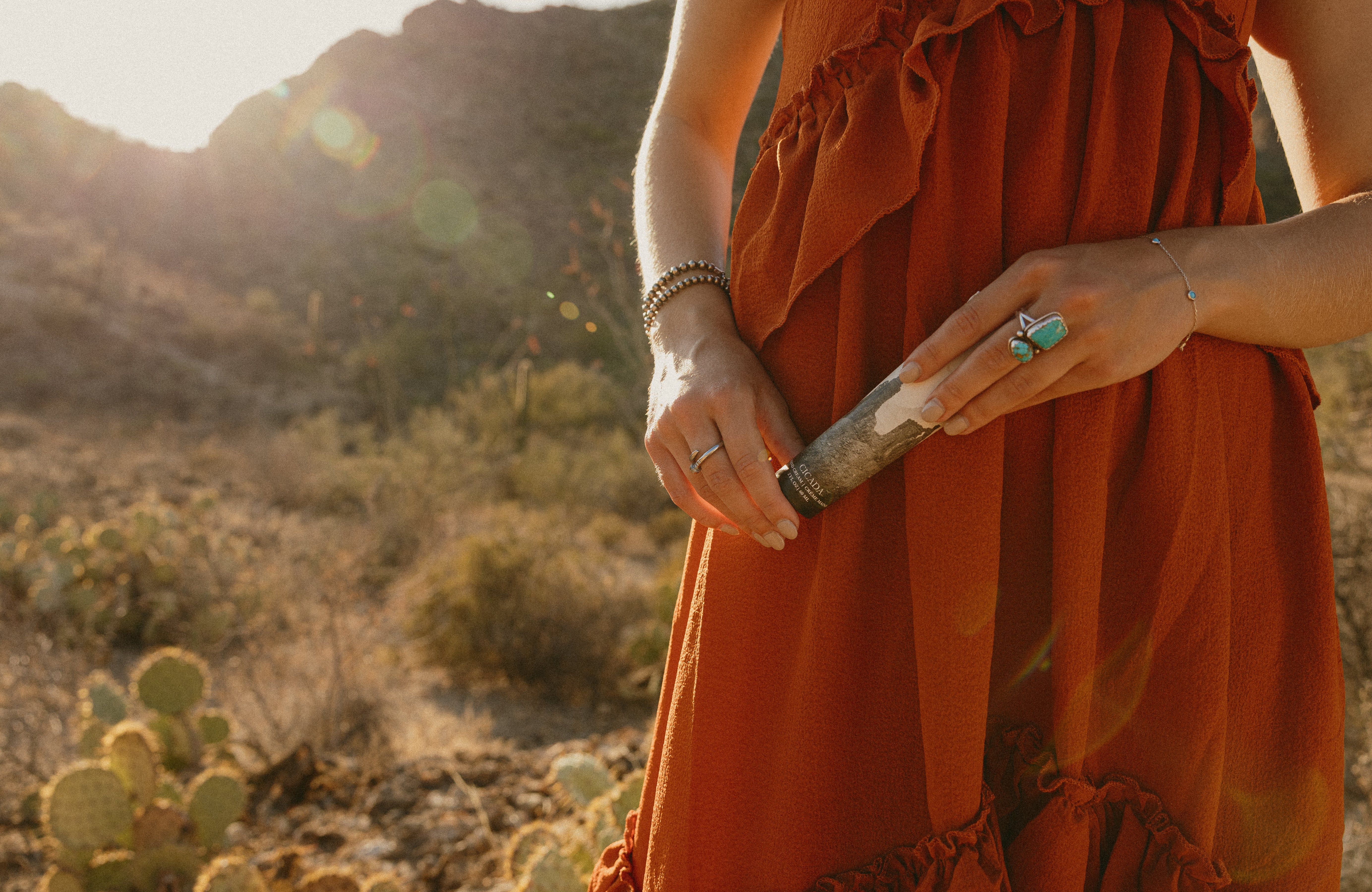 Woman in a red dress applying CICADA hand cream to her forearm while holding a CICADA cream tube, photographed outdoors in a desert setting.