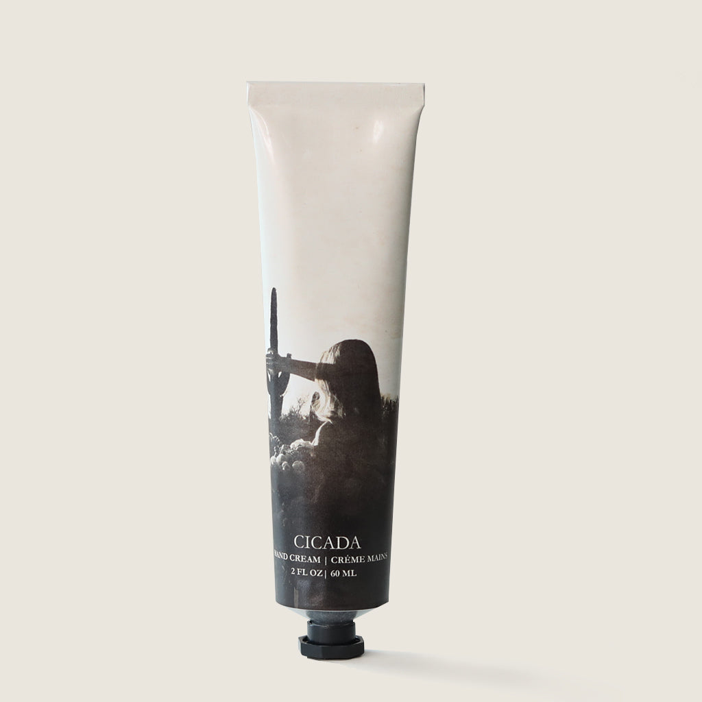 Product image of CICADA aluminum tube hand cream with a label showing a black and white photograph of a person standing in the desert. The top of the tube is white, and it has the text 'CICADA' and 'hand cream' in English and French as well as the product weight of '2 oz' or 60ml on it.  Image has light cream background. 