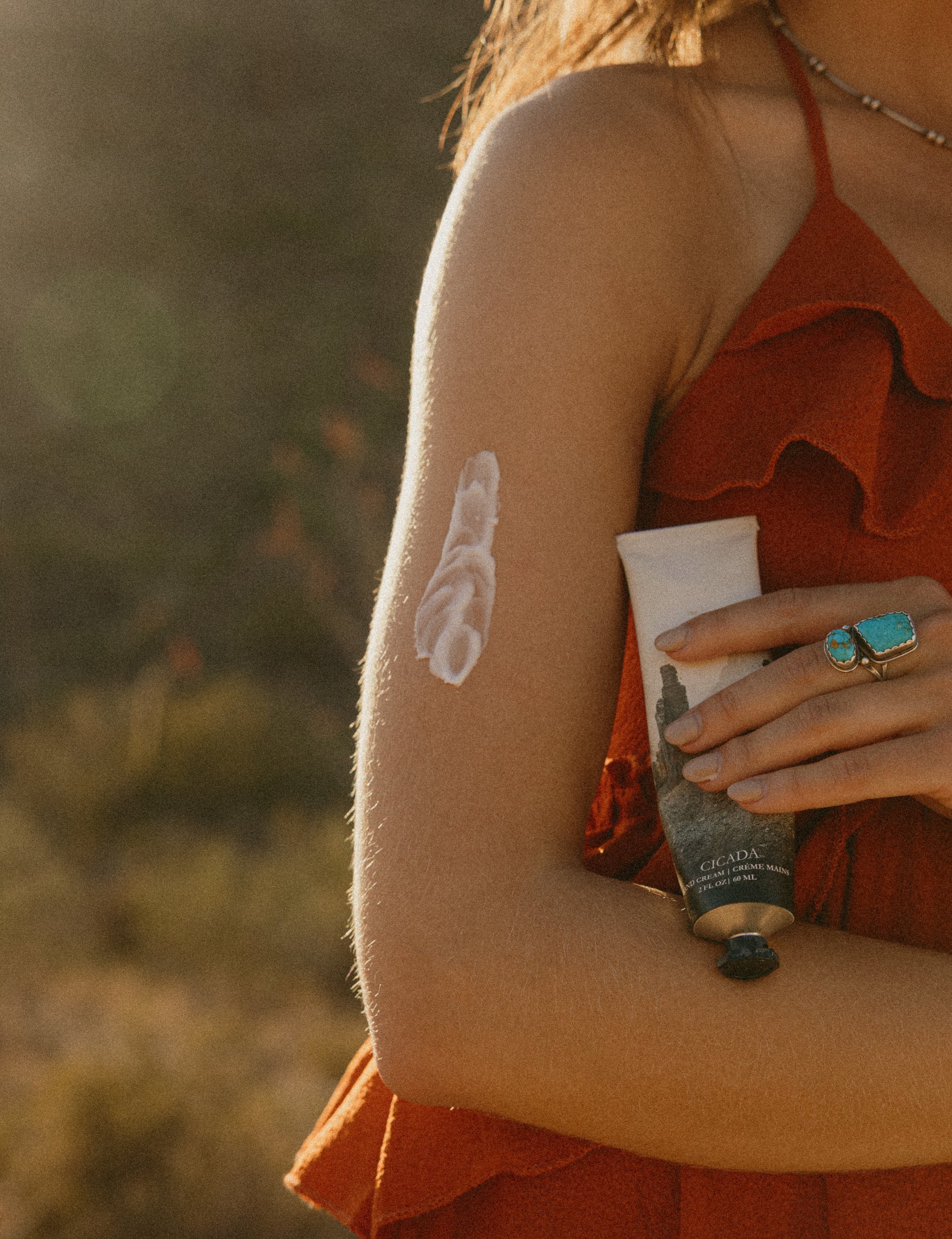 Lifestyle image with woman's arm with hand cream on it. Woman is holding CICADA hand cream with other hand. Woman is standing in the desert and wearing red dress. 