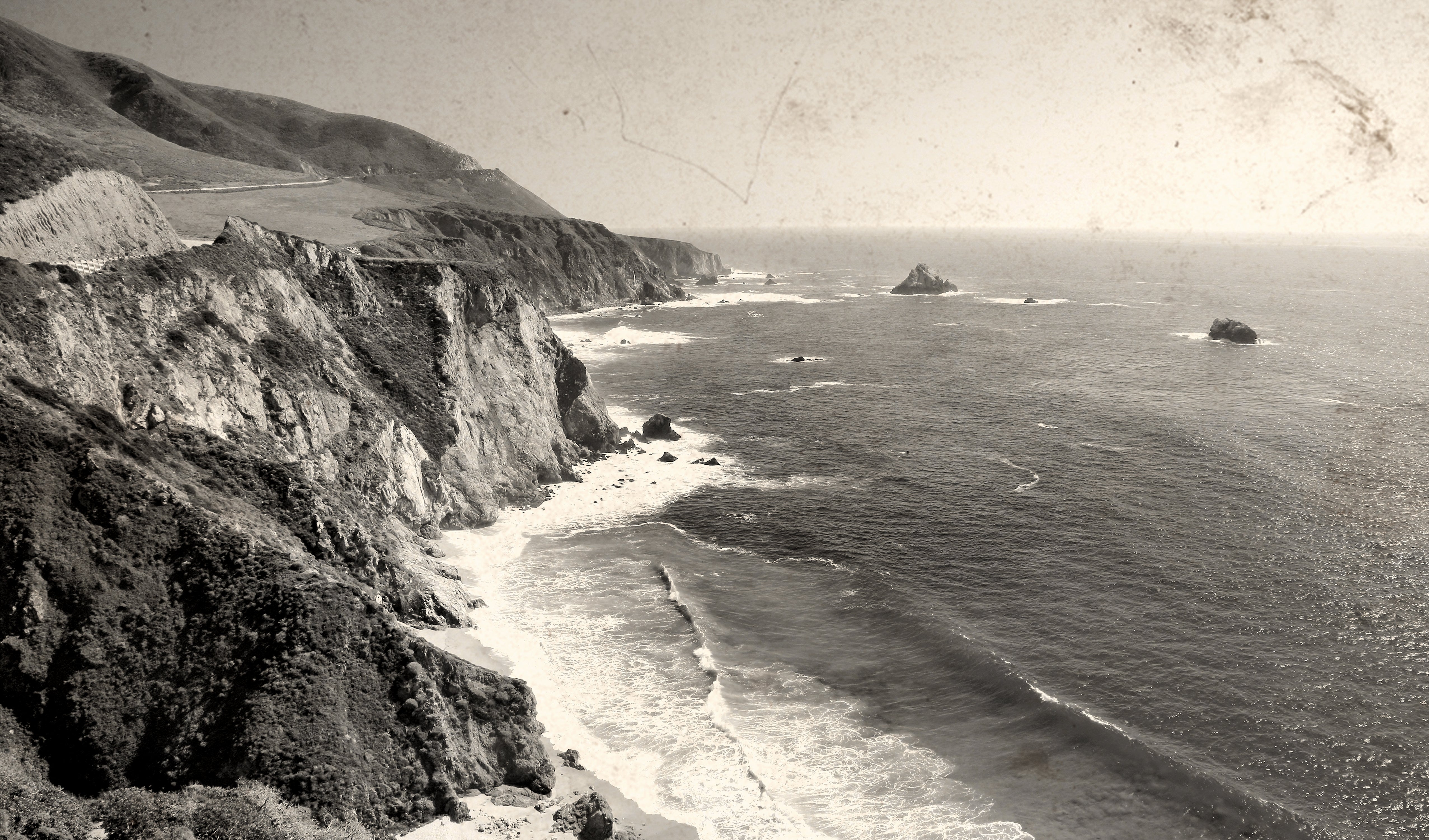 A vintage black and white photograph of a rugged coastal landscape, featuring steep cliffs dropping into the ocean with waves crashing along the shore and small rocky islands in the water.