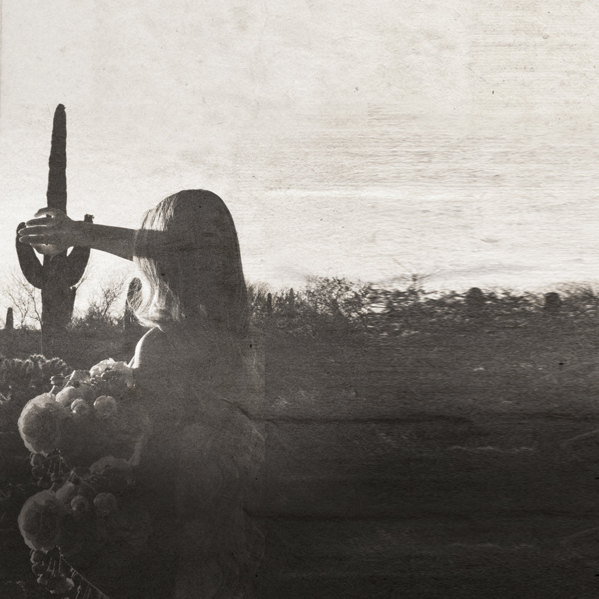 A sepia-toned artistic photograph showing a woman in profile holding a cactus outdoors in a desert landscape, with a large blurred foreground floral shape obscuring the lower left portion of the frame.