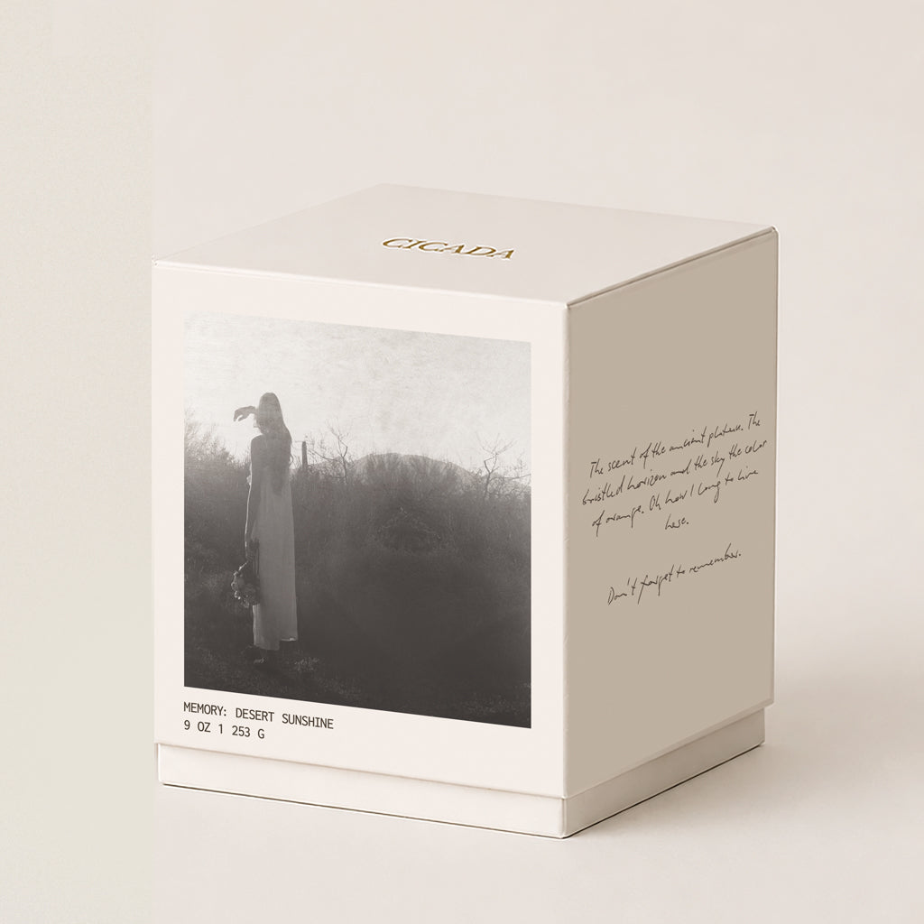 A cream 9oz candle box with a printed black-and-white photo of a woman standing outdoors, handwritten-type script on the side, and the brand name CICADA printed in gold at the top, shown on a warm off-white background.