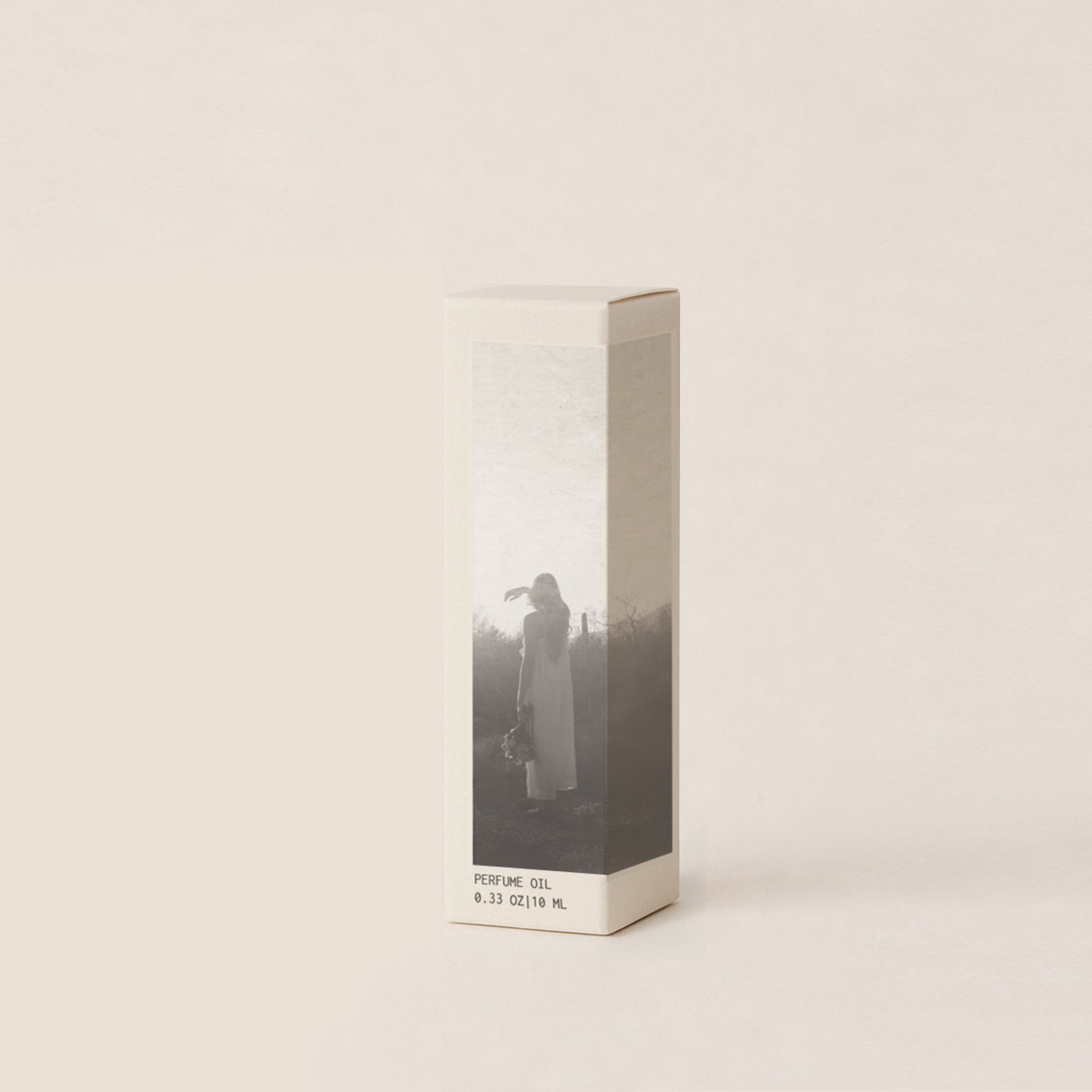A rectangular box for perfume oil, featuring a minimalist black-and-white photograph of a woman standing in a field, on a warm off-white background.