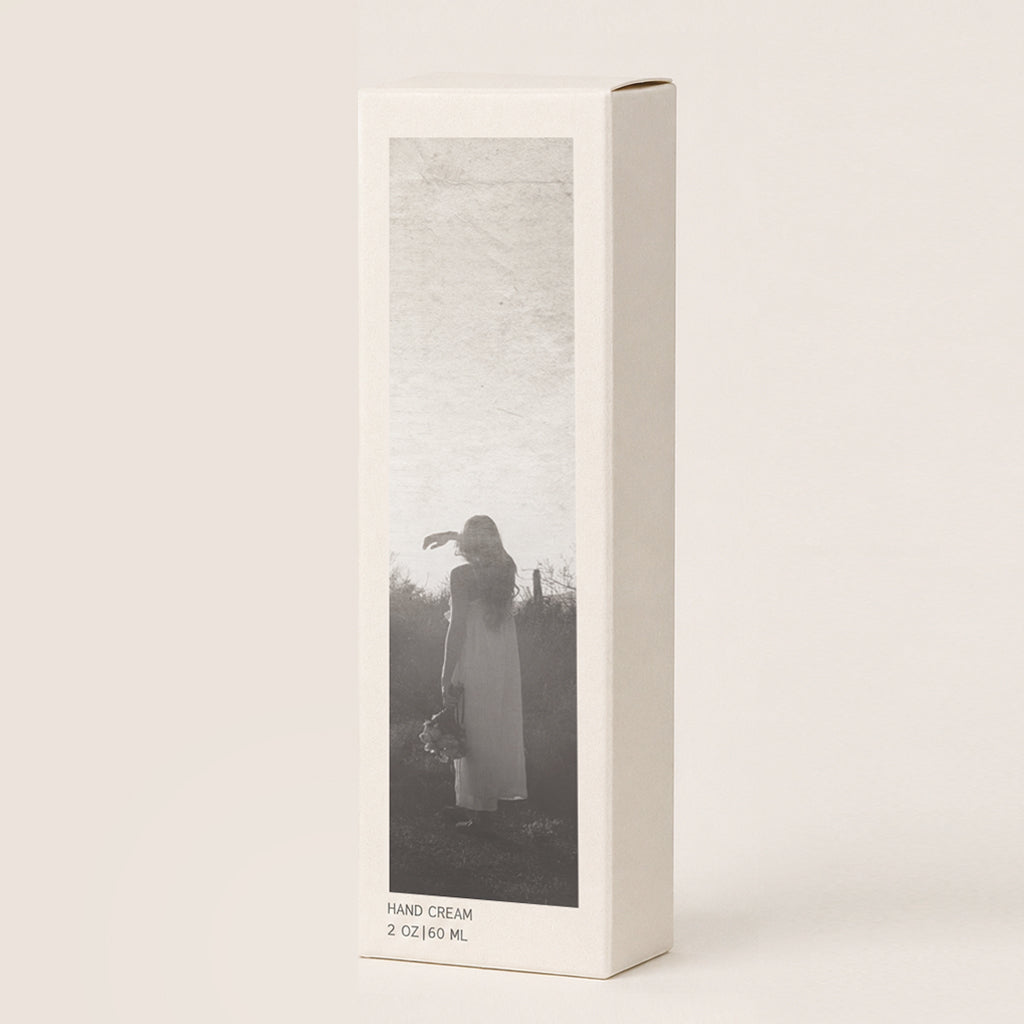 A tall, narrow hand cream box standing upright against a warm off-white background. The front of the box features a vertical black-and-white photograph of a woman in a long dress standing outdoors. 