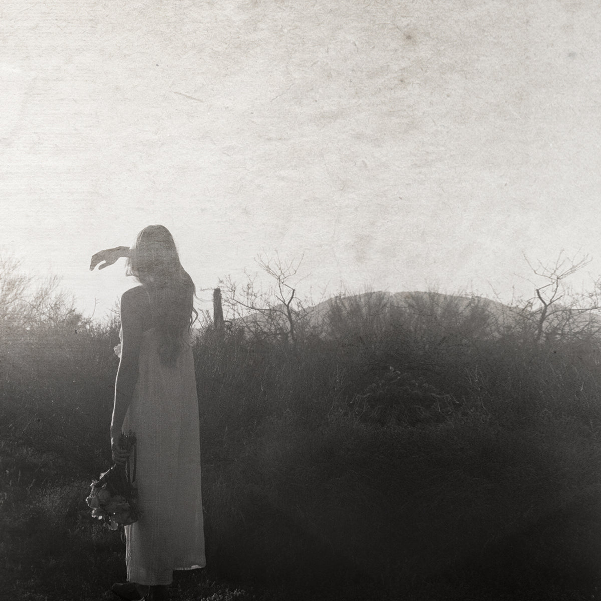 A monochrome, textured photograph of a woman in a long white dress standing outdoors in a field, holding a bouquet and looking toward the bright horizon.