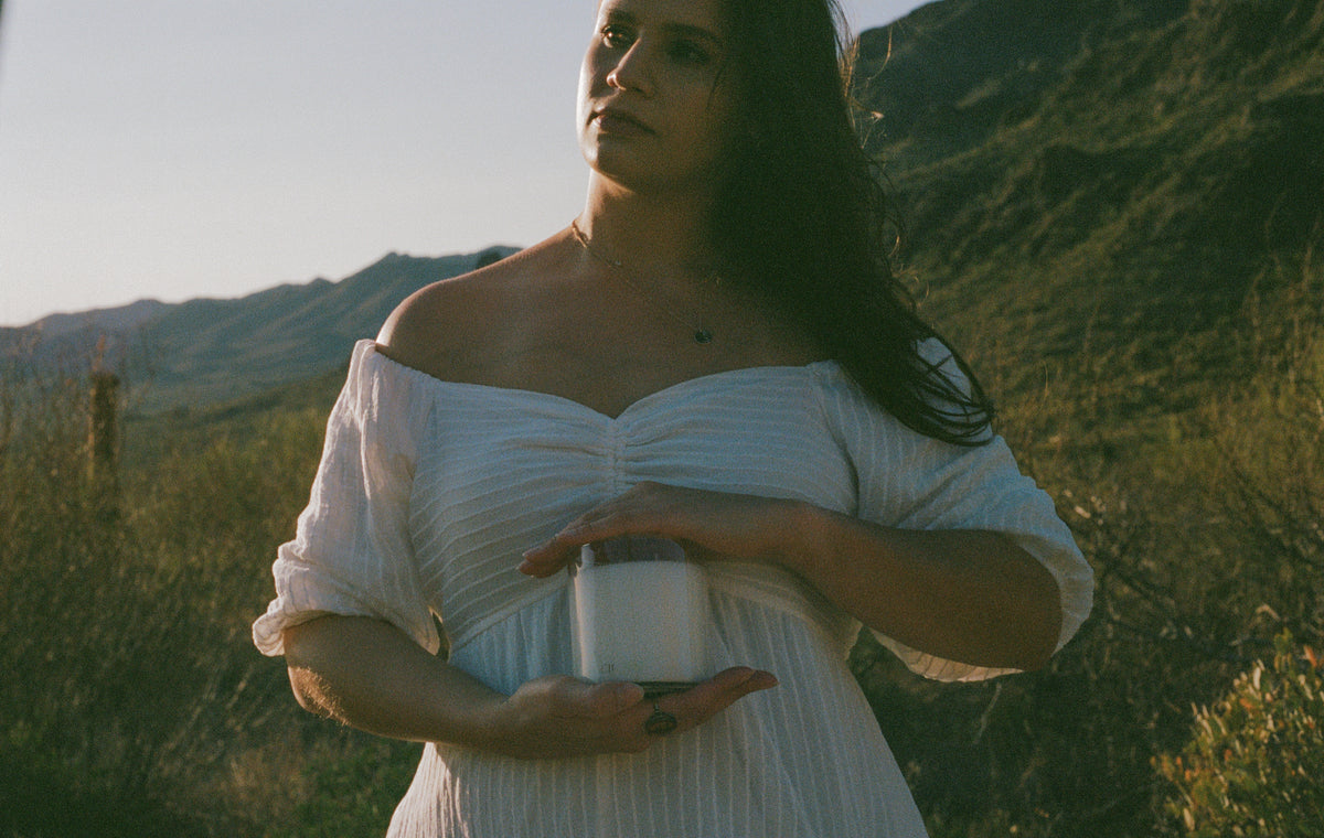 A woman wearing a white off-the-shoulder dress stands outdoors in a grassy, mountainous landscape at sunset, holding a white candle.