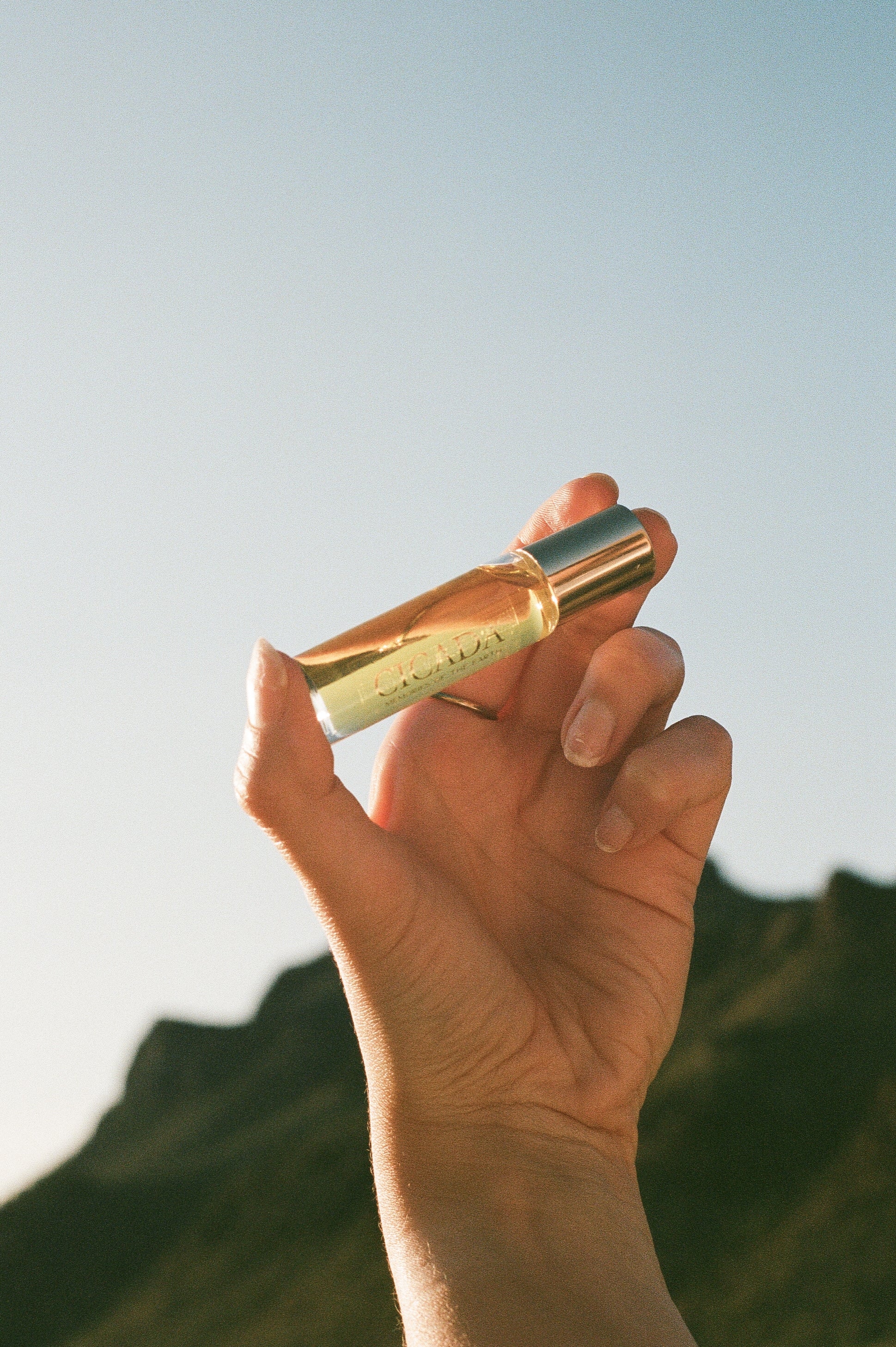 A hand holding a small glass perfume bottle with a metallic gold cap against a bright sky and blurred mountain background.