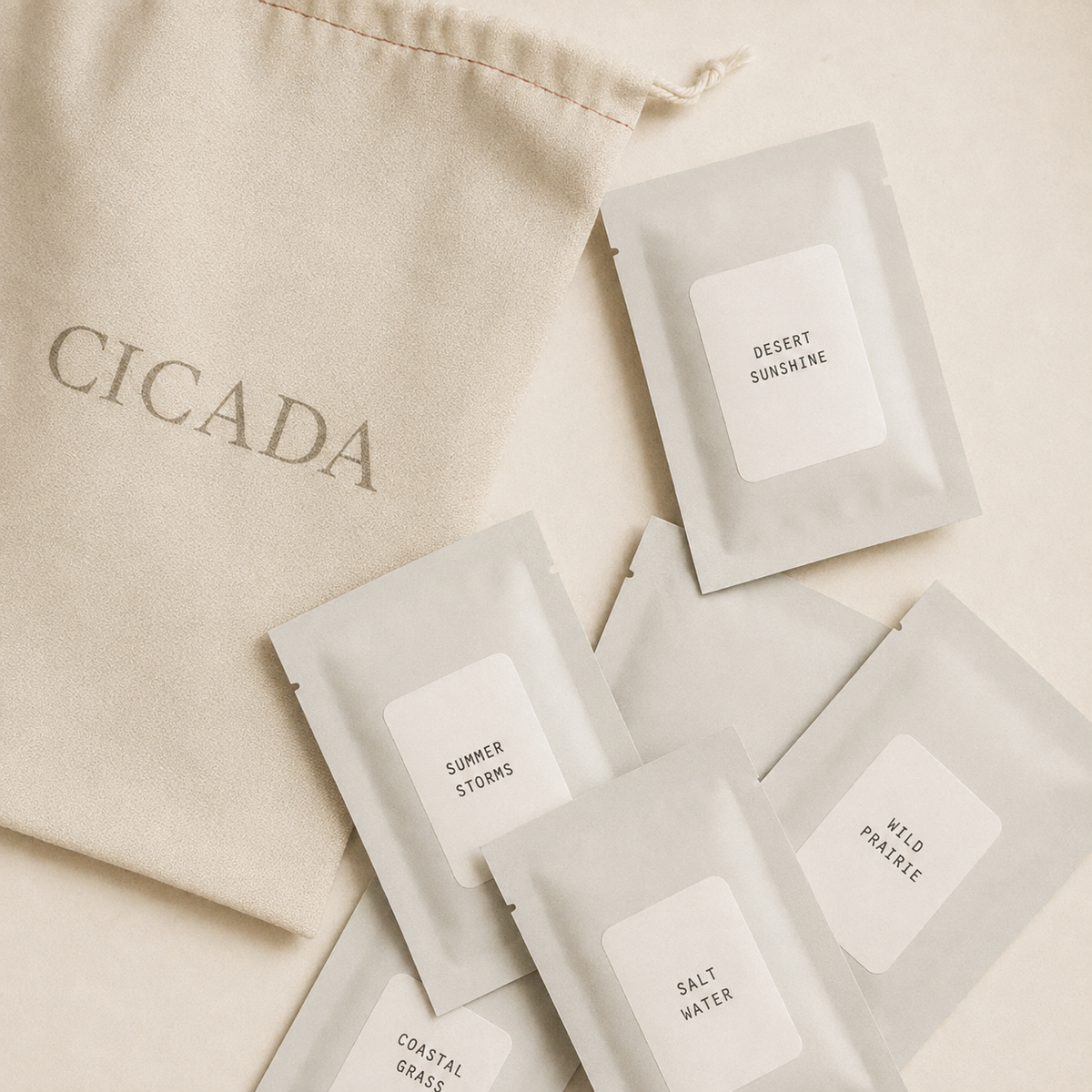 A beige drawstring pouch printed with the brand name CICADA is arranged on a light neutral surface with several small scent sampler packets. The packets have white labels reading Desert Sunshine, Summer Storms, Wild Prairie, Salt Water, and Coastal Grass.