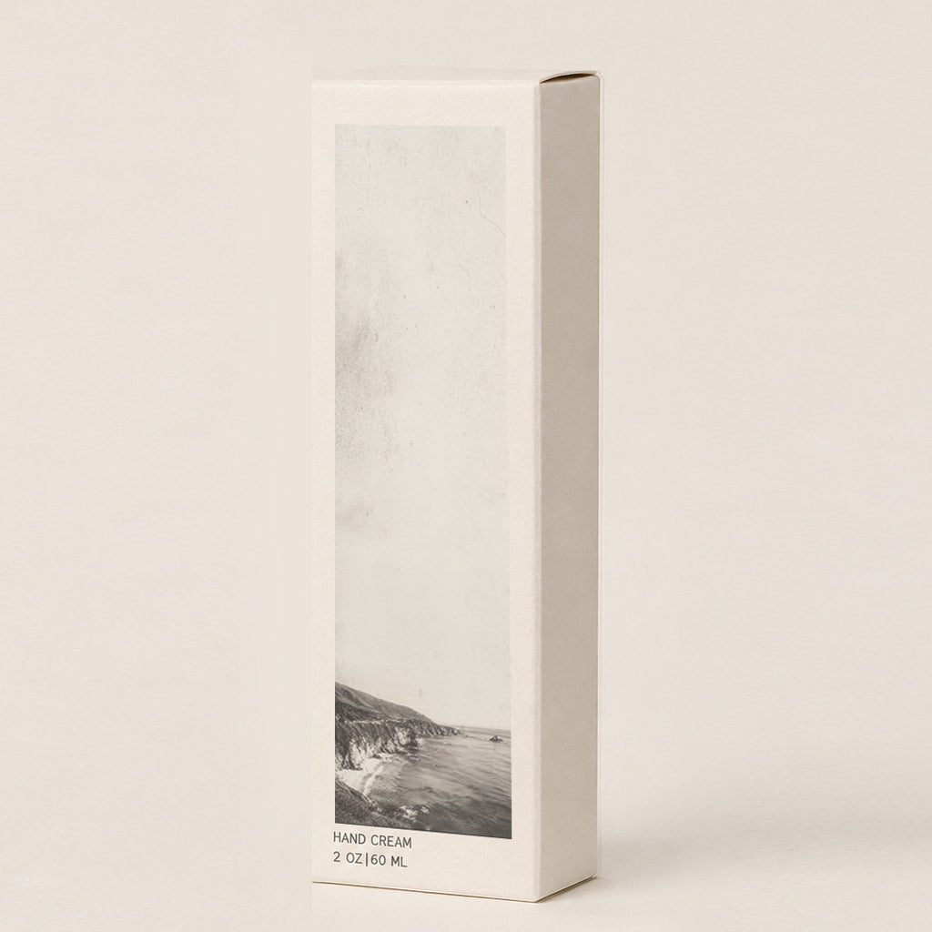 A tall, narrow cream colored rectangular box for hand cream, featuring a vertical black-and-white coastal landscape print on the front and the text 'HAND CREAM 2 OZ (60 ML)' near the bottom, photographed on a warm off-white background.