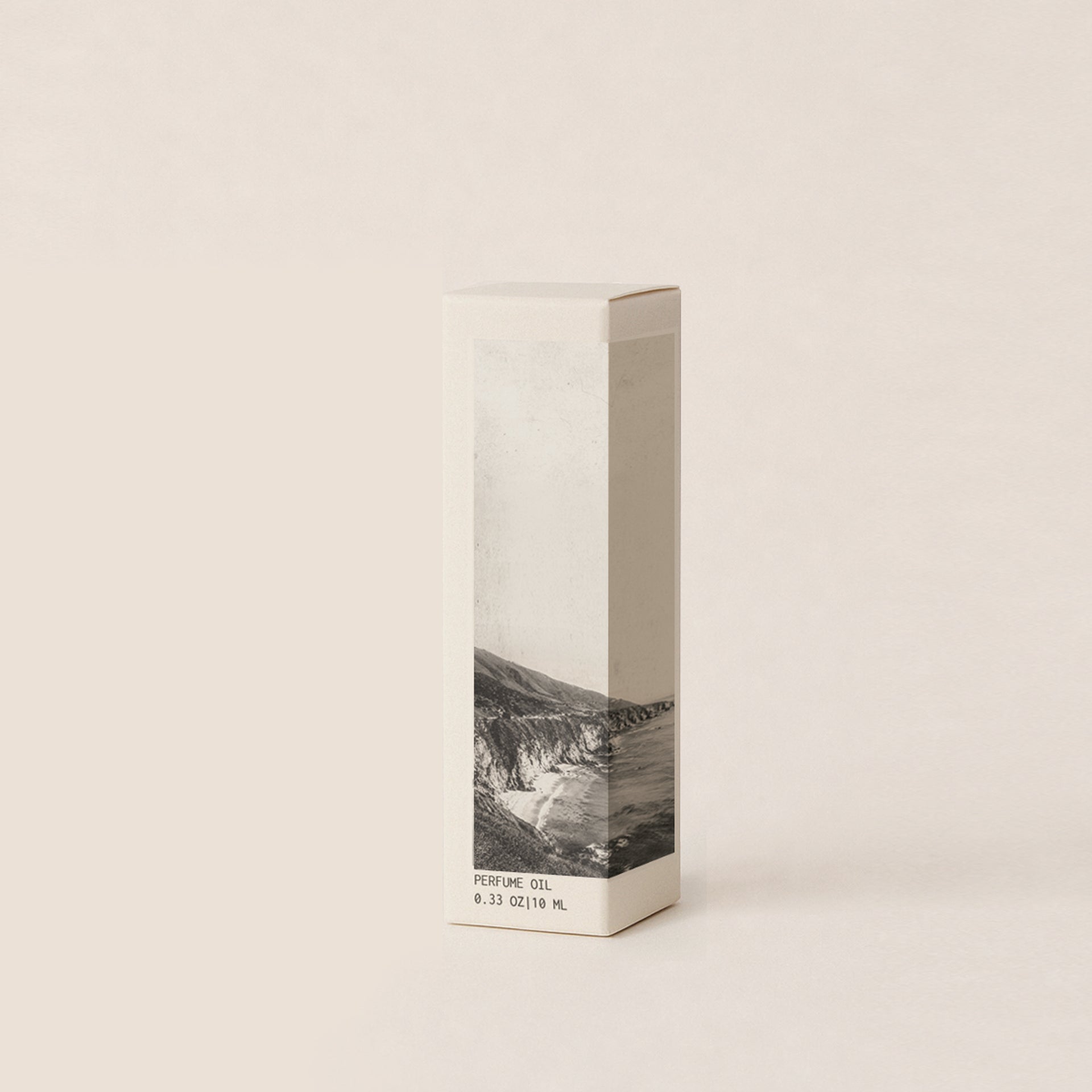 A rectangular perfume oil box with a minimalist design, featuring a black and white coastal landscape photograph on the lower half and small text reading 'PERFUME OIL' and '6033 OZ (10 mL)', set against a solid warm off-white background.