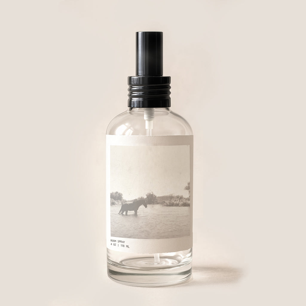 image of glass room spray bottle with black cap. Bottle has sepia archival image of horse in river on front label. 