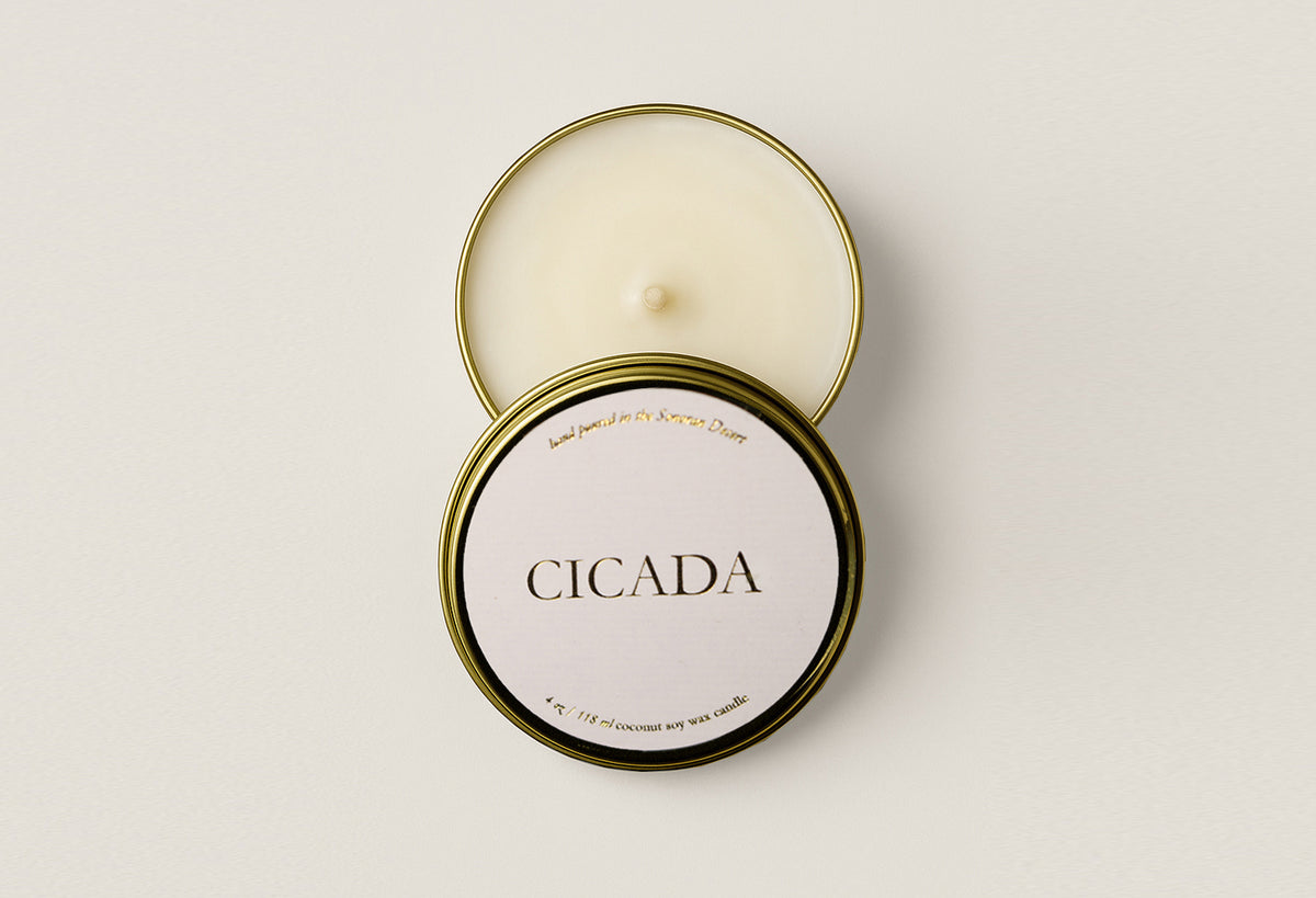 An open round metal candle tin showing cream-colored wax, with the lid placed on top. The front of the lid has a white label reading 'CICADA' in black serif text, with small gold text around the edge.